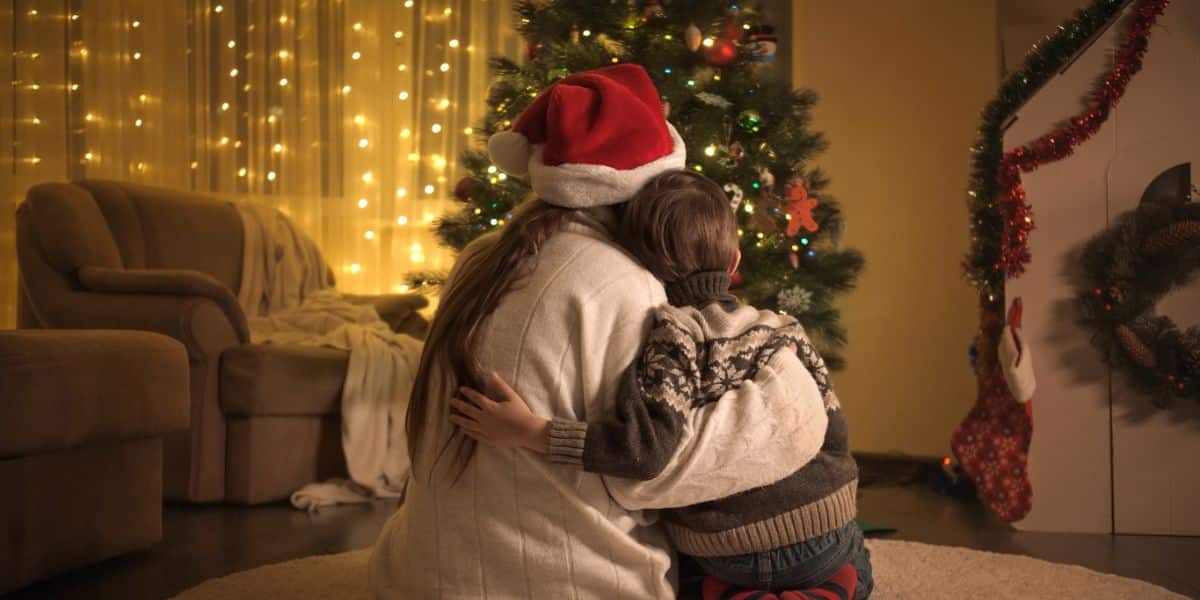 Mother in a Santa hat hugs her young son beside a lit Christmas tree at home during the Christmas holiday custody schedule